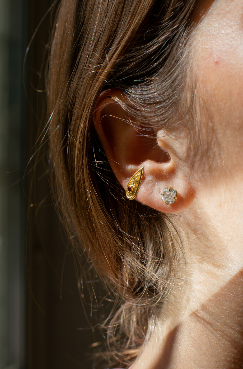 ear closeup on a gold teardrop-shaped earring set with a burgandy gemstone , and a gold & diamond stud earring