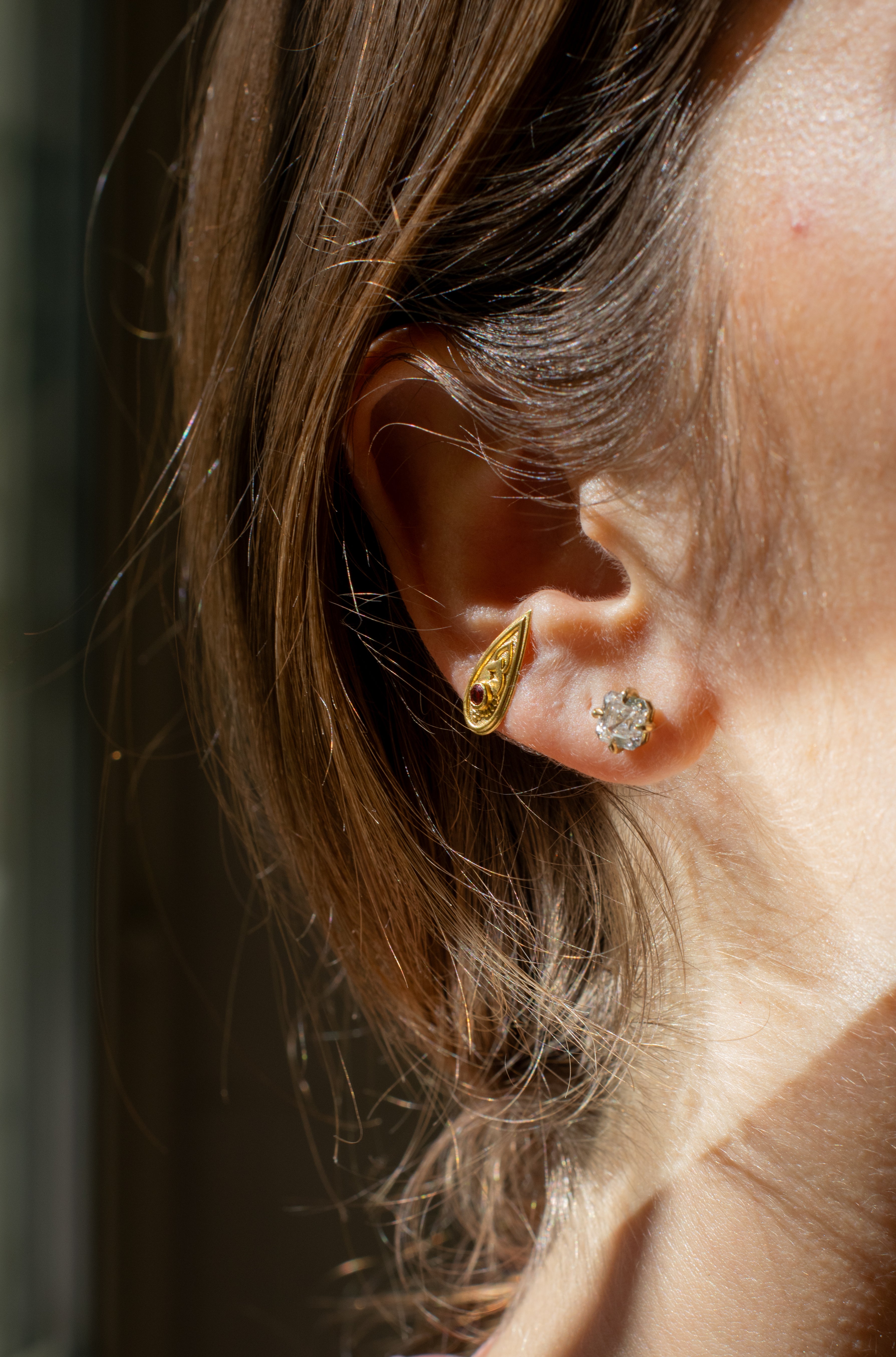 ear closeup on a gold teardrop-shaped earring set with a burgandy gemstone , and a gold & diamond stud earring